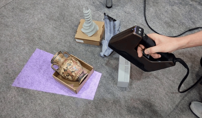 A handheld 3D scanner being used to digitize a Greek ceramic amphora vase and various 3D printed objects.