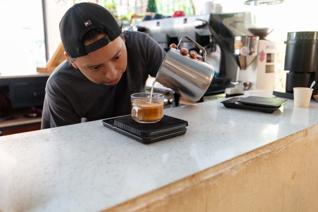 A barista pouring steamed milk into a glass of espresso to create a cappuccino on a coffee scale.