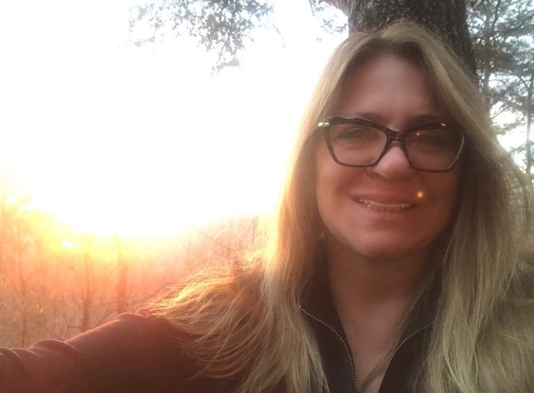 Smiling blonde woman with glasses taking a golden hour selfie during a scenic forest sunset.