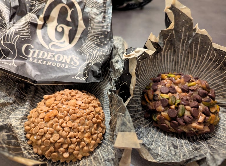 Two Gideon’s Bakehouse cookies on branded parchment, one with caramel chips and one with chocolate and pistachios.