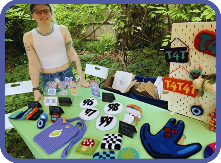 Artist selling handmade punch needle crafts and textile accessories at an outdoor market stall.