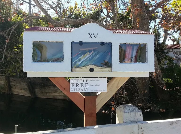 A white stucco Little Free Library shaped like a house with three glass window compartments filled with books.