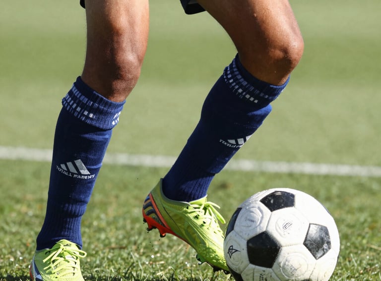 Close-up of a young soccer player's feet, diverse origin, performing a technical dribble with a modern soccer ball. European training facility background. The Total Football Academy logo is subtly placed on the player's sock. Bright daylight, dynamic motion blur.