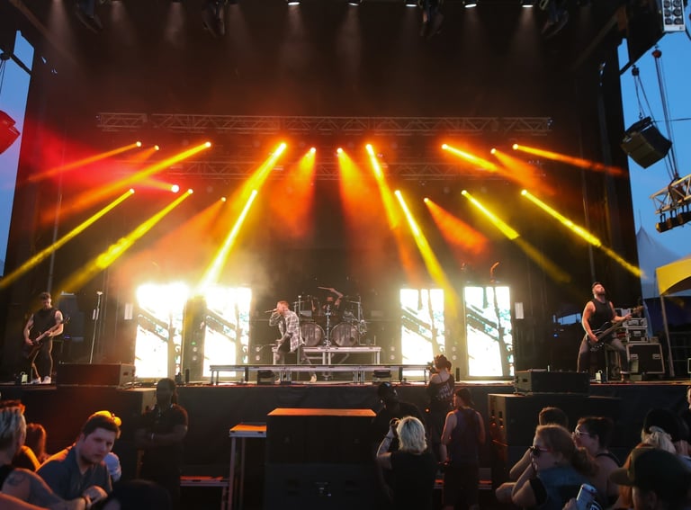 Live rock band performing on an outdoor stage with dramatic orange spotlights and a large crowd.