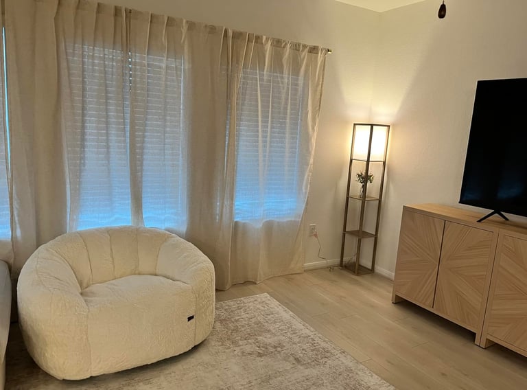 Cozy living room with a white sherpa bean bag chair, linen curtains, and a wooden TV stand.