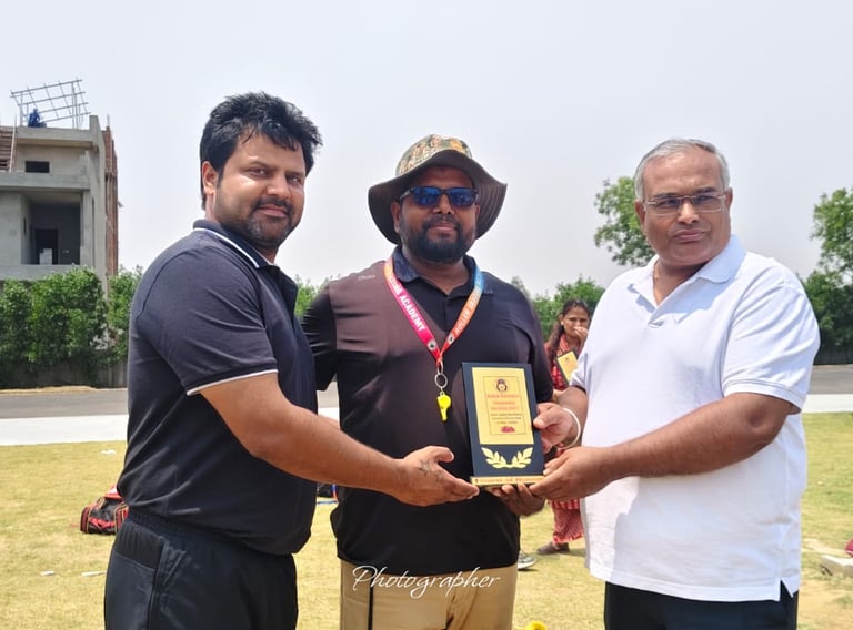 A man in a white polo shirt presents a sports award trophy to a coach on a sunny grass field.