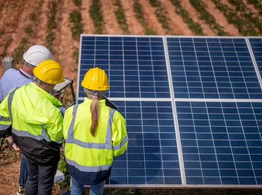 Fotografía de dos hombres y una mujer con cascos de protección color amarillo frente a paneles solar