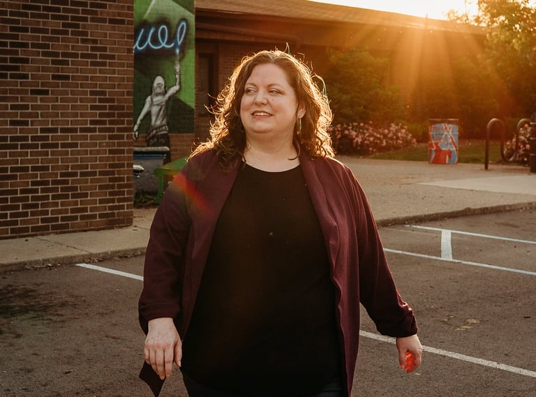 A woman walks through a paved parking lot during a golden hour sunset outside a brick building.