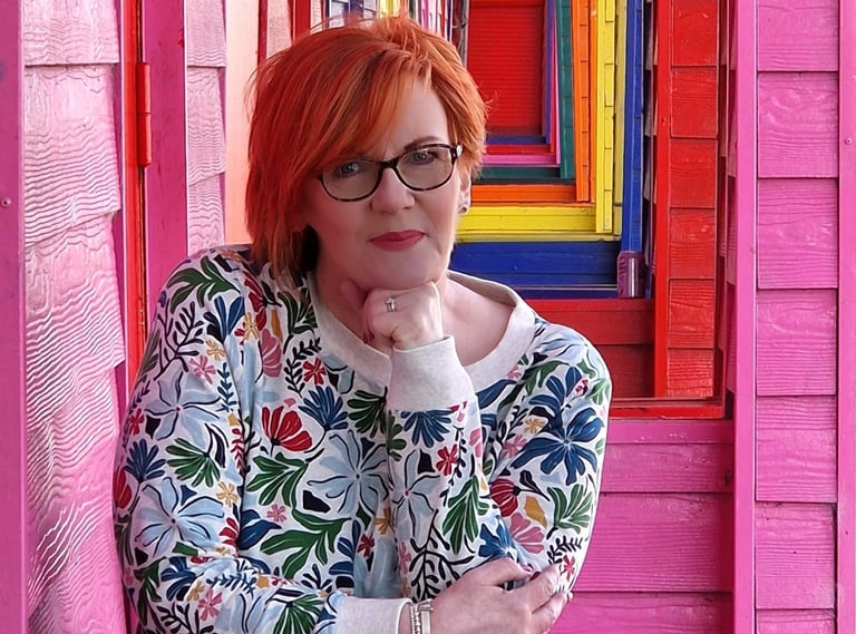 Dawn Vincent Celebrant leaning on a pink beach hut with a row of colourful huts in the background