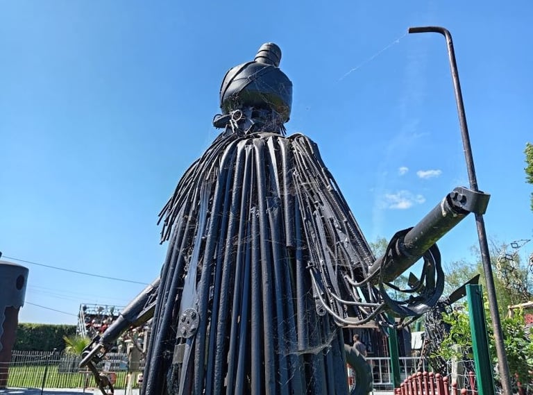 A tall black metal sculpture of a robed figure with a staff made from recycled pipes and scrap metal.