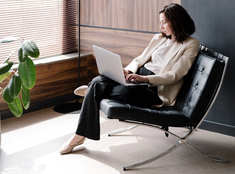 A business woman in a cream jacket and dark slacks,sitting on a black chair, working on a laptop.
