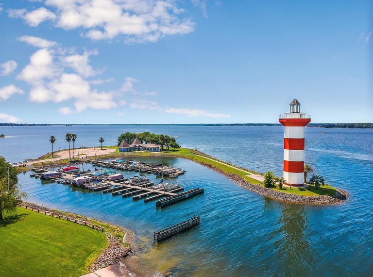 Aerial view of a red and white striped lighthouse at a boat marina on a sunny day with blue water.