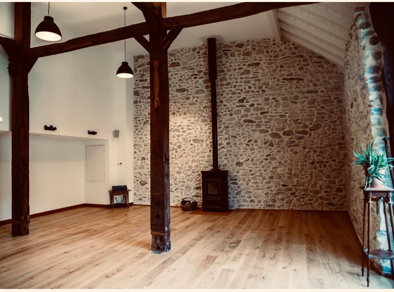 Rustic renovated barn interior featuring exposed stone walls, wood floors, and a modern black wood-burning stove.