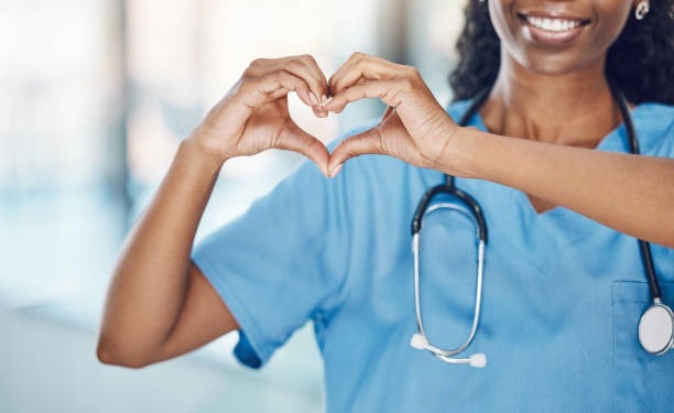 A smiling black woman in scrubs forms a heart shape with her hands, conveying warmth and compassion.