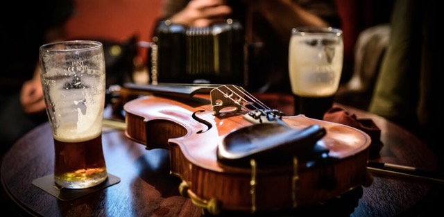 A wooden fiddle sits on a pub table alongside glasses of beer and an accordion.