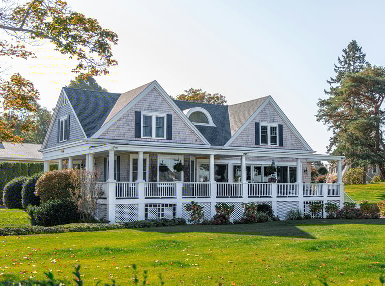 A luxury Cape Cod style shingle house featuring a large wrap-around porch and green lawn.