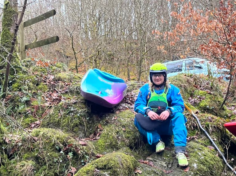 a woman in a dry suit with a white water kayak on a mossy hill 