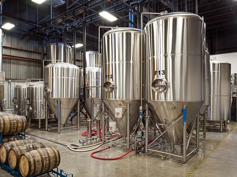 Large stainless steel conical fermenters and oak aging barrels inside a modern craft brewery facility.