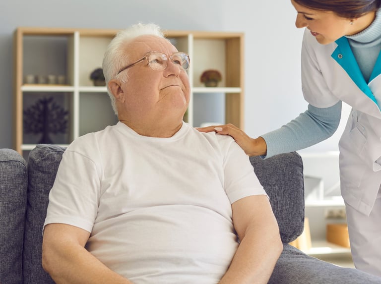 un homme âgée assis sur un fauteuil la main d'une soignante sur l'épaule gauche.