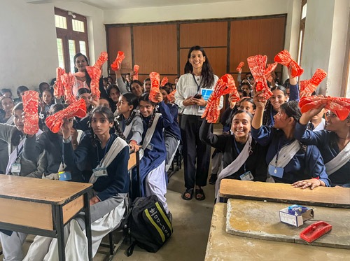 About 20 girls in classroom holding up napkin packs. with Pooja.