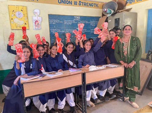 About 12 girls in classroom holding up napkin packs.