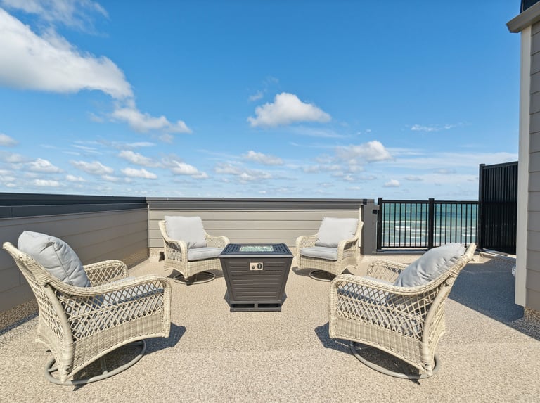 Wicker patio chairs surrounding a modern fire pit on a rooftop deck with beachfront views.
