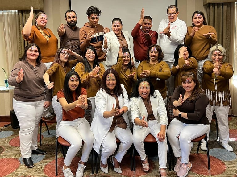 Diverse group of people in white pants and brown tops posing with forehead jewels for a team photo.