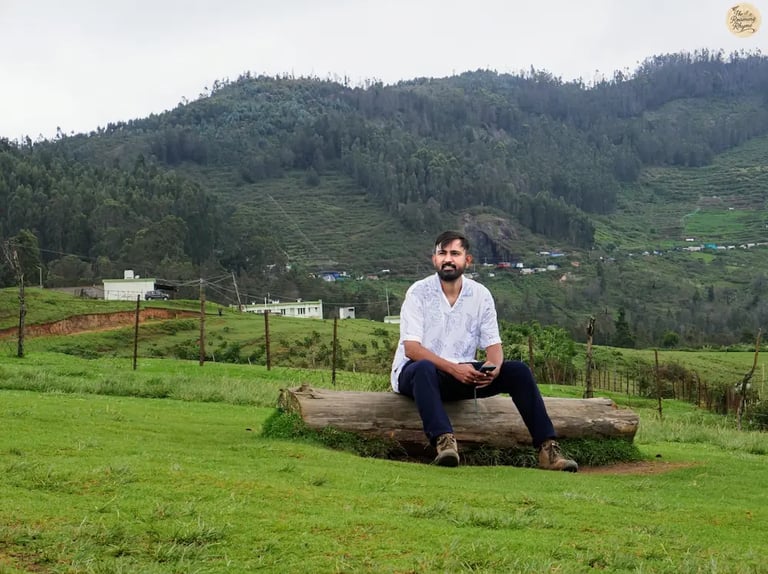 Traveler sitting on a tree trunk at Mannavanur Sheep Farm, surrounded by rolling meadows and hills.