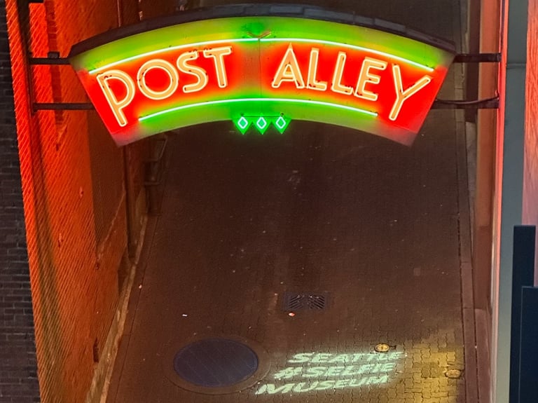 A neon sign saying post Allie with a holographic projection of Seattle #selfie museum underneath