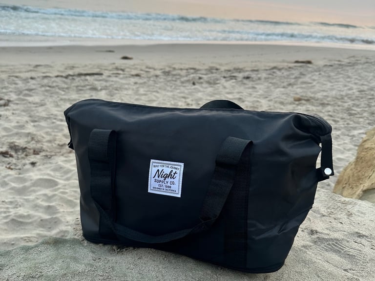Black Night Supply Co. duffel bag on beach sand with sunset sky in background