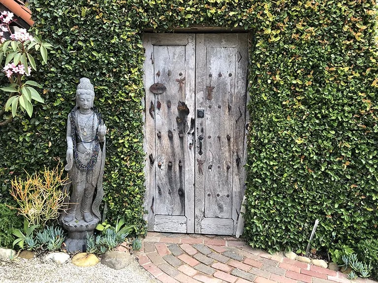 Wood buddha statue by weathered wood gate and vine-covered wall.