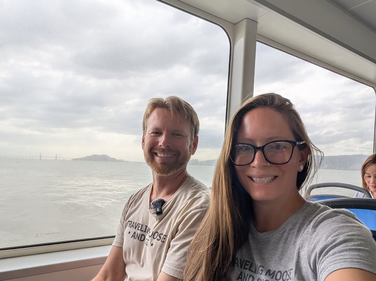 A smiling couple sits on a ferry boat to Penang with a scenic ocean view through the window.