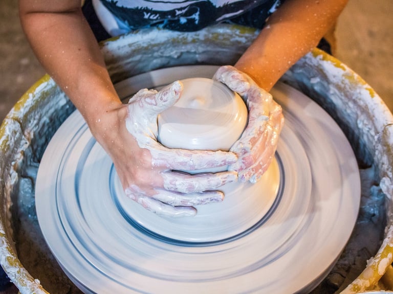 An individual's hands throwing a mound of clay on a potter's wheel.
