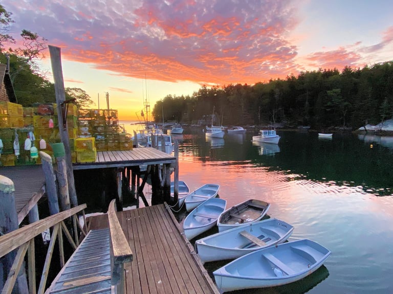 Fishing boats at their moorings beside a wooden dock at dawn, pink and orange sky reflected in still water.