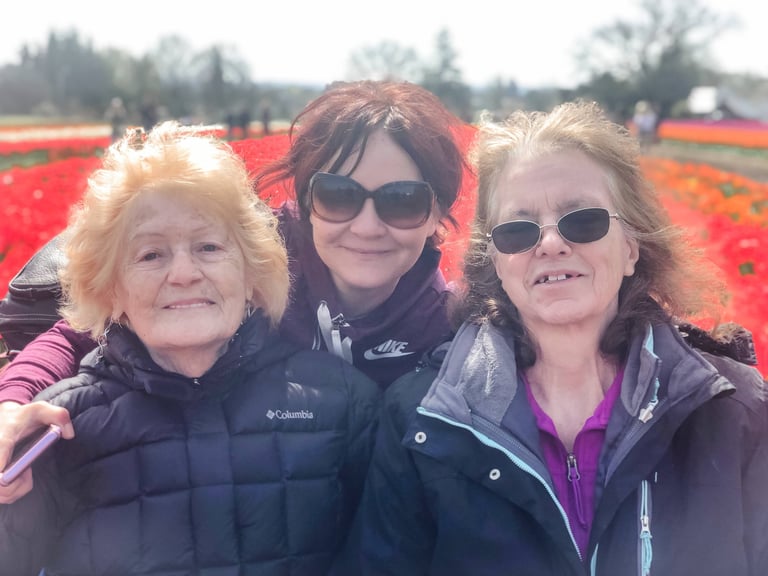 Three happy women wearing jackets and sunglasses smiling in a vibrant red tulip field.