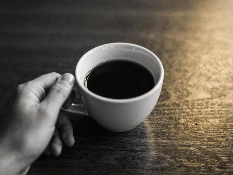 Close-up of a hand holding a cup of strong coffee on a polished dark wood surface, subtle golden light, black and white filter, 35mm film.