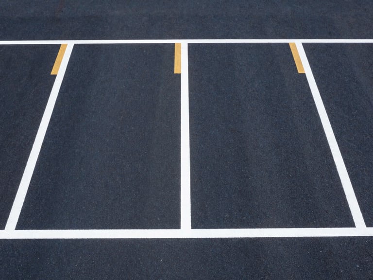 Symmetrical overhead drone shot of freshly applied, razor-sharp white and safety-gold parking lines on dark asphalt. Bright midday sun casts high-contrast, crisp shadows, emphasizing the flawless layout alignment.