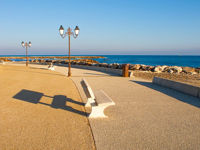 Empty stone benches and vintage street lamps along a paved seaside promenade with blue ocean views.