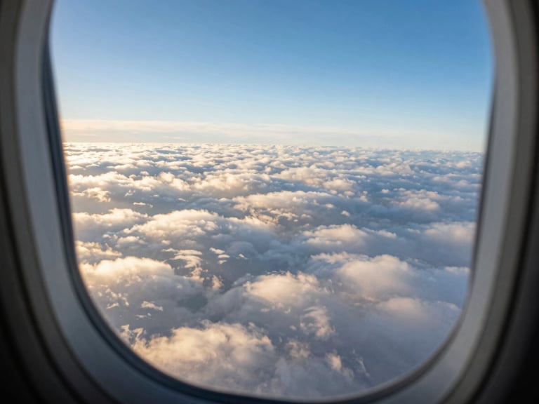 Clean aircraft window overlooking a vast expanse of clouds during golden hour, first-person perspective, wide-angle cinematic lens, rich saturated blues and whites.