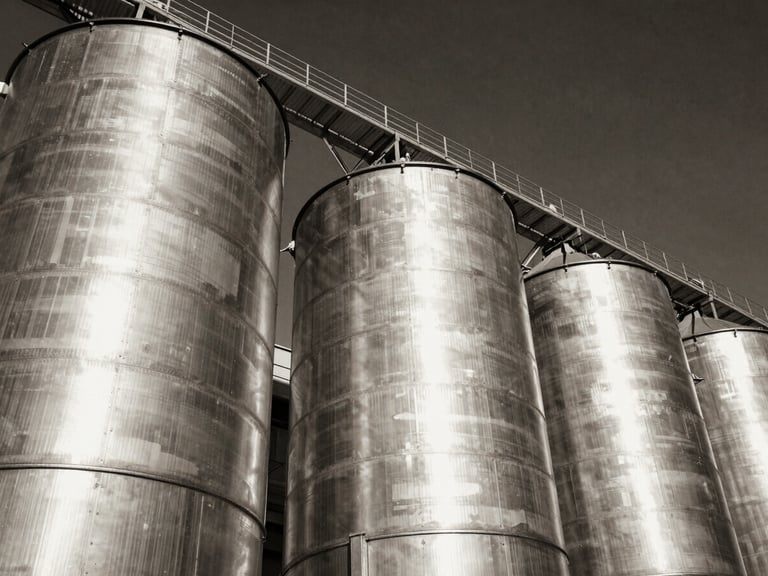 Low-angle heroic perspective of massive galvanized steel grain silos under dramatic side-lighting, high contrast black and white tones