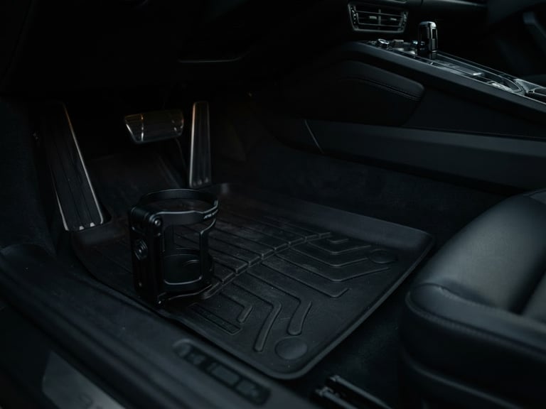 Low-angle close-up shot of an Apex Mounts cup holder securely clipped to a heavy-duty black floor mat in a Porsche 911 footwell, moody directional cabin light, deep shadows, matte textures, sharp focus, 50mm lens. The footwell is clean and minimalist, showing pedal box and part of the transmission tunnel.