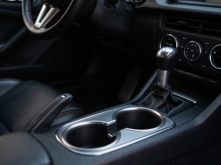 Close-up of Apex Mounts cup holder installed low in a Mazda MX-5 Miata footwell, showing ample clearance around the manual shifter, moody cabin light, deep shadows, matte textures.