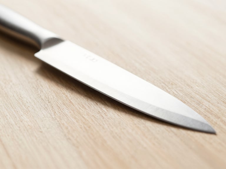 Tight macro shot of a sharp steel chef's knife resting on a clean wooden surface, bright diffused natural daylight, shallow depth of field, warm neutral tones.