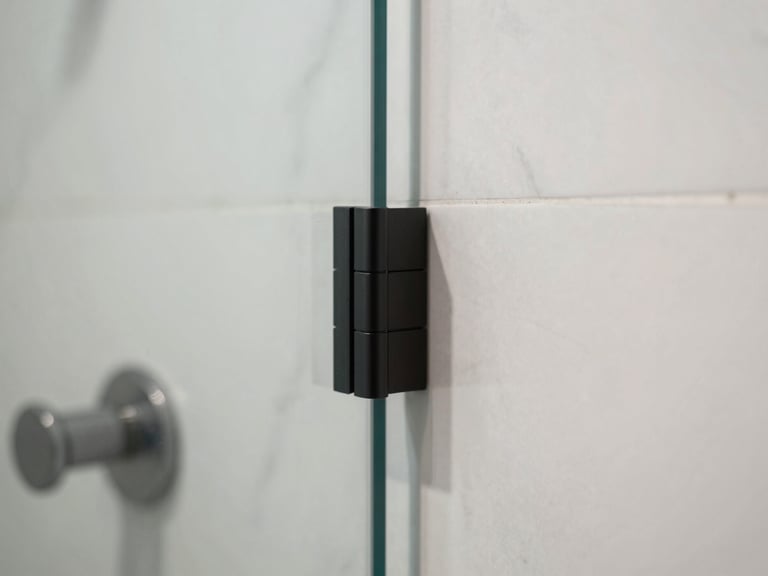 Close-up of a frameless glass shower enclosure joint, matte black hardware profile meeting a clean marble wall, natural diffused daylight, sharp focus on the mitred edge