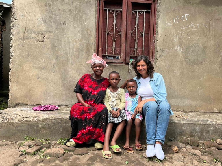 A smiling group of two women and two children sitting outside a house in an African village.