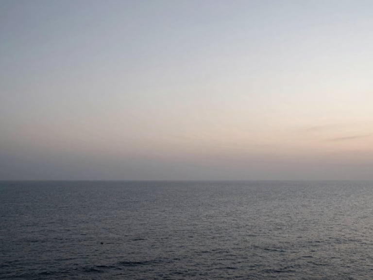 Wide panoramic shot of an ocean horizon at dawn, soft desaturated morning light, overcast sky, vast negative space, calm coastal atmosphere.