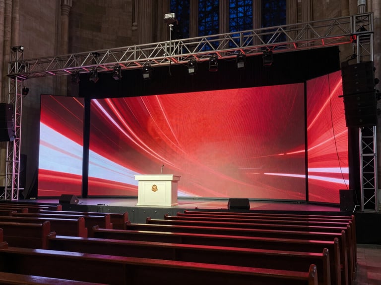 Wide environmental shot of a church sanctuary at night, a large LED wall glowing with deep red and white stage graphics behind the pulpit, tungsten key light washing the front rows, intricate truss rigging visible in the upper frame, cables dressed precisely, cinematic and controlled atmosphere