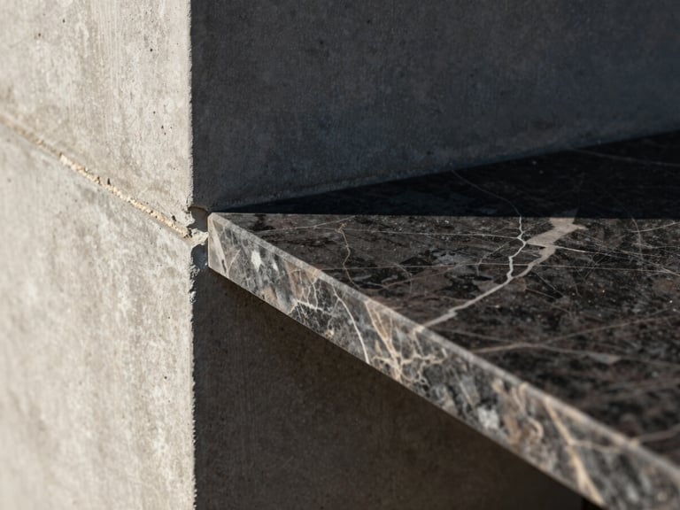 Close-up detail crop of a dark marble slab meeting a raw concrete wall, sharp sunlight highlighting the precise joint line, landscape orientation.