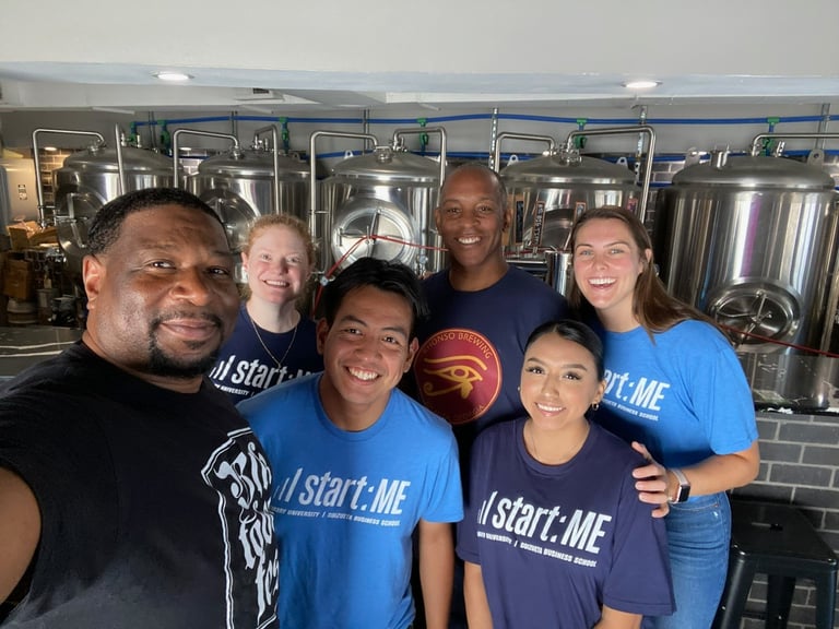 A group of diverse entrepreneurs smiling in a brewery with large stainless steel fermentation tanks.