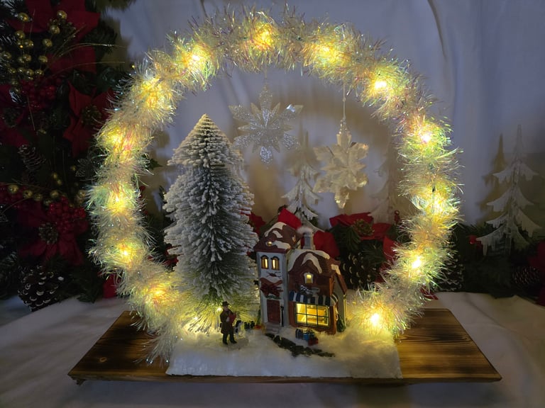 Illuminated Christmas village scene with a tinsel wreath, snowy miniature tree, and cozy light-up house.